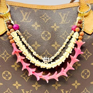 May include: A brown monogram canvas tote bag featuring tan leather trim and handles. A decorative multi-strand beaded charm hangs from the front, showcasing pink coral-like branches, cream-colored carved beads, and small brown wooden spheres.
