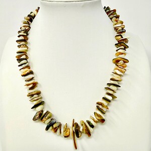 May include: A necklace composed of numerous irregular, polished chip beads in shades of cream, tan, brown, and dark green. The beads are strung together in a single strand, featuring a slightly elongated, vertical bead at the center bottom.