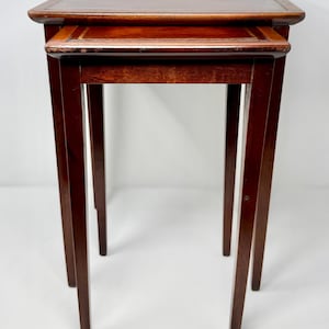 May include: Set of two nesting tables. The tables are made of dark brown wood with a glossy finish. The tops are rectangular with rounded corners. The legs are slender and straight. The smaller table fits neatly under the larger one.