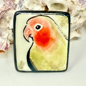 May include: A square ceramic tile featuring a hand-painted illustration of a lovebird with a bright orange face and a pale green body. The bird is depicted in profile against a light cream background, framed by a thick, dark, hand-painted border.
