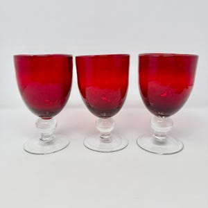 May include: Three red wine glasses with a clear, round base and stem. The bowl of each glass is a deep red color, with a slightly lighter shade towards the bottom. The glasses are arranged in a row against a white background.