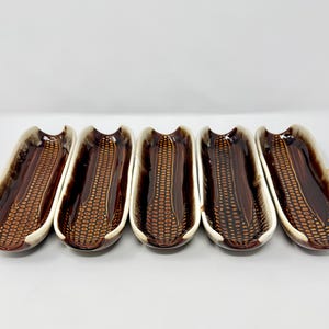 May include: Five ceramic corn-shaped serving dishes. Each dish is brown with a textured pattern resembling corn kernels, and a cream-colored rim. The dishes are arranged in a row against a white background.