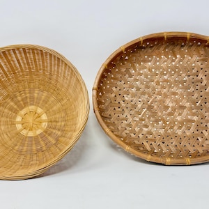 May include: Two round woven baskets made of light brown natural material. The basket on the left features a deep, bowl-like shape with vertical radiating slats, while the basket on the right is a shallow, flat tray with a hexagonal weave pattern.