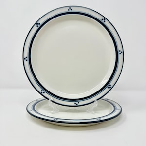 May include: Two white ceramic plates with a navy blue border and dotted accents. The plates are round and have a simple, elegant design. One plate is stacked on top of another, showcasing the design.