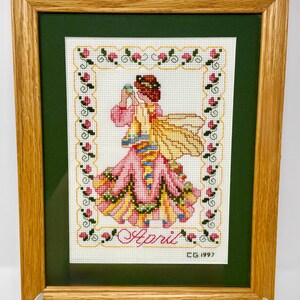 May include: Framed cross-stitch artwork featuring a woman in a pink dress with yellow wings, holding a small object. The artwork is framed in a wooden frame with a green mat. The word "April" is stitched at the bottom.