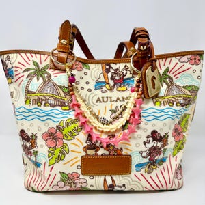 May include: A cream-colored tote bag featuring a colorful tropical print with Mickey and Minnie Mouse, palm trees, and the text AULANI. The bag has brown leather handles and a decorative beaded charm with pink and white shell-like accents.