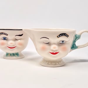 May include: Two cream-colored ceramic face mugs. One is a small cup with a winking face, a bow tie, and the word "Baileys". The other is a creamer with a handle, a green bow, and the word "Baileys". Both have rosy cheeks and red lips.