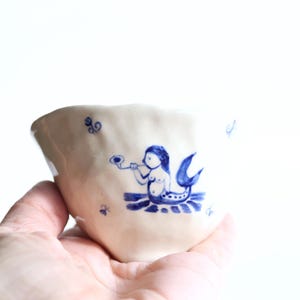 May include: A handmade ceramic cup with a whimsical design. The off-white cup features a blue illustration of a mermaid playing a horn. The cup is held in a hand against a white background. The cup has a unique, irregular shape.