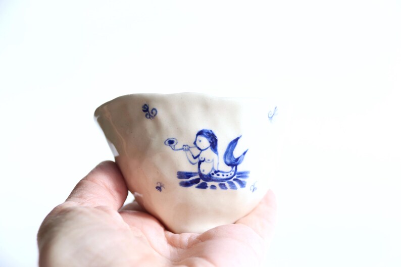 May include: A handmade ceramic cup with a whimsical design. The off-white cup features a blue illustration of a mermaid playing a horn. The cup is held in a hand against a white background. The cup has a unique, irregular shape.