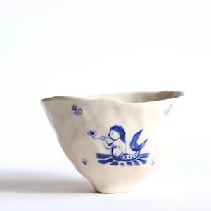 May include: A white ceramic bowl with a blue mermaid illustration. The mermaid is blowing a horn. The bowl has an irregular, wavy rim and is decorated with small blue swirls.
