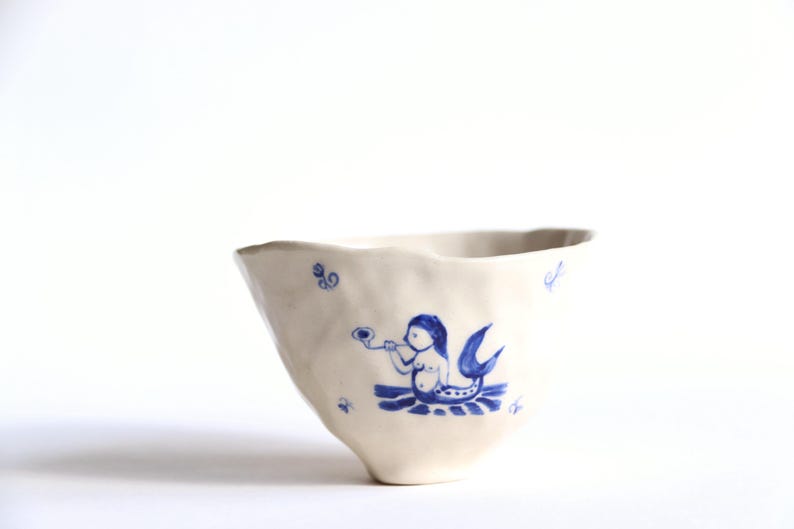 May include: A white ceramic bowl with a blue mermaid illustration. The mermaid is blowing a horn. The bowl has an irregular, wavy rim and is decorated with small blue swirls.