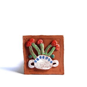 May include: A terracotta tile with a raised design of a white ceramic pot with red and green flowers. The tile has a brown border.