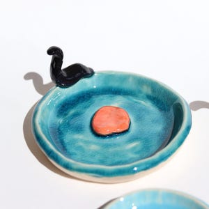 Può includere: Un piattino rotondo in ceramica con smalto turchese lucido, caratterizzato da una piccola creatura nera scolpita appollaiata sul bordo e un singolo accento circolare arancione piatto centrato sulla base interna della ciotola.