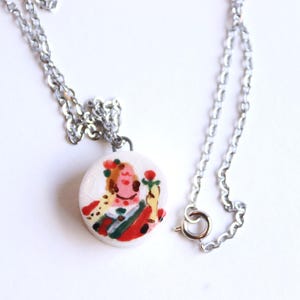 Puede incluir: Un collar plateado con un colgante redondo. El colgante muestra una ilustración pintada a mano de una figura en rojo, verde y amarillo. El colgante es blanco con una textura agrietada. El collar tiene una cadena y un cierre sencillos.