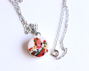 Op de afbeelding: Een zilverkleurige ketting met een ronde hanger. De hanger toont een handgeschilderde illustratie van een figuur in rood, groen en geel. De hanger is wit met een gebarsten textuur. De ketting heeft een eenvoudige ketting en sluiting.