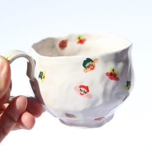 Peut inclure: Une tasse en céramique blanche avec une forme irrégulière façonnée à la main et une anse incurvée. Les surfaces extérieure et intérieure sont décorées de petites illustrations peintes à la main représentant des visages aux cheveux verts, rouges et jaunes.