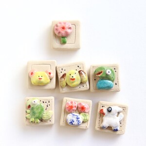 May include: A collection of seven handmade ceramic tiles, each featuring a unique, colorful, and whimsical design. The tiles depict a flower, a koala, a dog, a lizard, and other creatures. The tiles are square and have a textured, off-white background.
