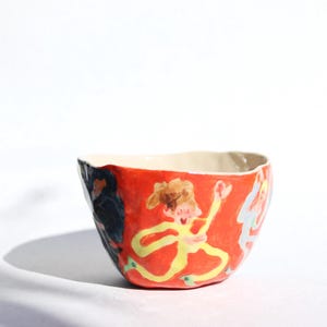 Puede incluir: Un pequeño cuenco de cerámica rojo con un diseño pintado a mano de fantasía con figuras en amarillo, azul y negro. El cuenco tiene un borde irregular, ligeramente desigual.