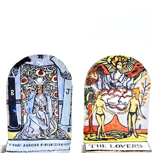 Peut inclure: Deux cartes de tarot en céramique, l'une représentant La Grande Prêtresse et l'autre Les Amoureux. La carte de la Grande Prêtresse est bleue et blanche avec une femme assise sur un trône. La carte des Amoureux est orange et jaune avec un couple debout devant un arbre.