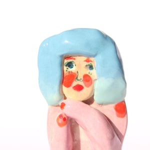 May include: A small ceramic figurine featuring a figure with bright blue hair, a pale face, and prominent red cheeks and lips. The figure wears a light pink garment with small red dots on the shoulders and arms, rendered in a smooth, glossy finish.