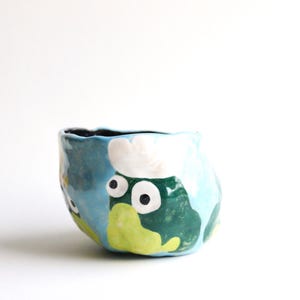 Könnte beinhalten: Eine blaue Keramikschüssel mit einem skurrilen Design, das einen grünen Frosch mit weißen Augen und einem weißen Hut zeigt. Die Schüssel hat eine strukturierte, unebene Oberfläche.