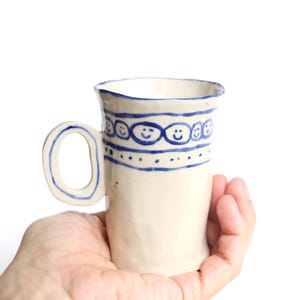 May include: A handmade ceramic mug with a white base and a blue smiley face design. The mug has a unique handle and is held in a person's hand. The mug is approximately 13 cm tall.