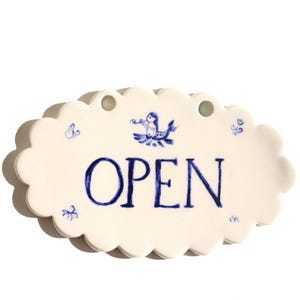 Puede incluir: Letrero de cerámica color crema con borde festoneado, con la palabra "OPEN" en escritura azul. El letrero está decorado con detalles florales azules y una ilustración central de una criatura mítica.
