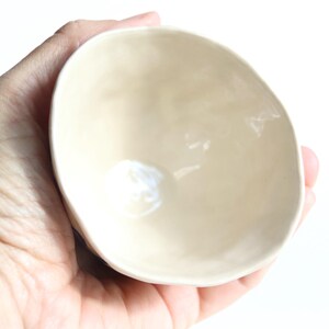 May include: A small, off-white ceramic bowl with a smooth, glossy finish. The bowl has an irregular, organic shape and is held in a hand against a white background. The interior of the bowl reflects light, highlighting its curved form.