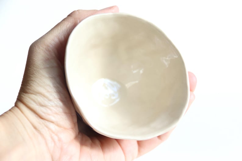 May include: A small, off-white ceramic bowl with a smooth, glossy finish. The bowl has an irregular, organic shape and is held in a hand against a white background. The interior of the bowl reflects light, highlighting its curved form.