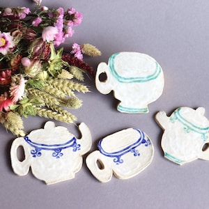 May include: Four ceramic teacups and teapots in white with blue and green painted details. The teacups and teapots are arranged on a gray surface.