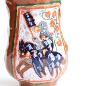 May include: A brown ceramic vase with a white and blue painted design of a knight riding a horse. The knight is wearing a blue and white suit of armor and a helmet with a plume. The horse is brown with white markings. The vase is decorated with orange flowers and green leaves.