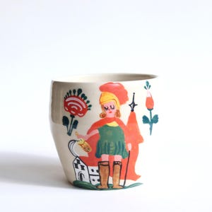 Puede incluir: Taza de cerámica color crema con una ilustración caprichosa. El diseño presenta una figura con un vestido verde y una capa roja, sosteniendo una lanza. Elementos florales y arquitectónicos rodean la figura, sobre un fondo blanco.