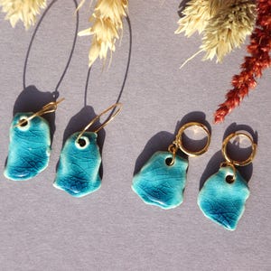 Puede incluir: Dos pares de pendientes de cerámica turquesa con aros dorados. Los pendientes tienen una forma irregular y un esmalte craquelado. Flores secas en el fondo, proyectando sombras sobre la superficie gris.