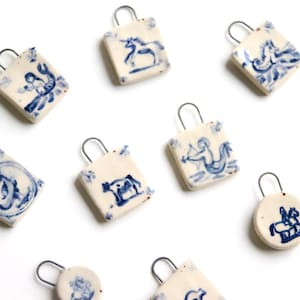 Könnte beinhalten: Zehn Keramik-Anhänger mit blauen und weißen Malereien. Die Anhänger sind in Form von kleinen Einkaufstaschen und Kreisen gestaltet. Jeder Anhänger zeigt ein anderes Design, darunter ein Einhorn, eine Meerjungfrau, eine Kuh und ein Pferd.