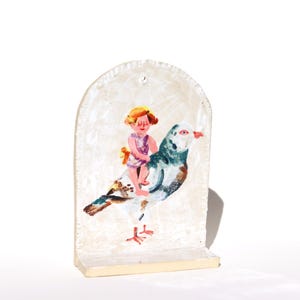 May include: A ceramic sculpture depicting a child riding a pigeon. The artwork uses a soft colour palette, with the child in a purple outfit and the bird in shades of blue, brown, and white. The sculpture has a textured, off-white background and a small base.