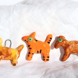 Puede incluir: Tres figuras de animales de cerámica: una criatura amarilla y marrón con forma de dinosaurio, un gato naranja con rayas negras y una cabra marrón. Cada uno tiene ojos verdes o rojos. Las figuras están sobre una tela blanca.