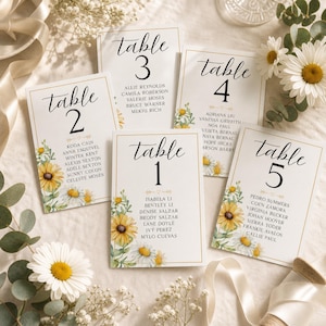 Puede incluir: Cinco tarjetas de mesa rectangulares blancas para bodas con bordes dorados, cada una con la palabra table seguida de un número del uno al cinco, una lista de nombres e ilustraciones en acuarela de girasoles amarillos y margaritas blancas.