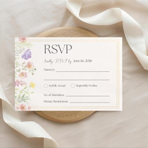 Puede incluir: Tarjeta RSVP crema con detalles florales. La tarjeta presenta la palabra "RSVP" y campos para nombres, aceptación y restricciones dietéticas. El diseño incluye flores de acuarela en rosa, morado y amarillo.