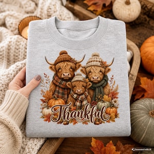 May include: A light gray crewneck sweatshirt featuring a graphic of three Highland cows wearing knit hats and plaid scarves. The cows are surrounded by autumn leaves and pumpkins, with the word Thankful written in brown script below them.