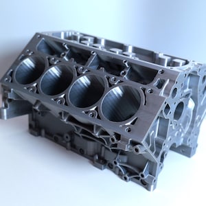 May include: A silver-colored metal V8 engine block featuring four visible cylindrical bores on the top surface, intricate cast structural webbing, and multiple bolt holes along the perimeter of the engine casing for mechanical assembly.