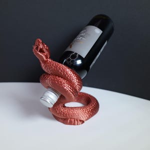May include: A metallic copper-coloured snake-shaped wine bottle holder with a textured scale pattern. The coiled snake body supports a dark glass wine bottle with a silver screw cap, angled upwards with the snake head raised and mouth open.
