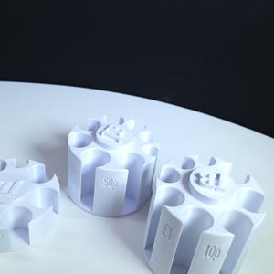 May include: Three white cylindrical coin sorters with multiple vertical slots. Each unit features embossed text labels including 1p, 10p, 50p, and £1, along with a central circular logo containing the letters TB. The items are arranged in a cluster.