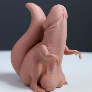May include: A matte, light brown, 3D-printed figurine featuring a creature with a large, curved tail, small arms, and a phallic-shaped head and torso. The object has a textured surface and sits in an upright position with visible anatomical details.