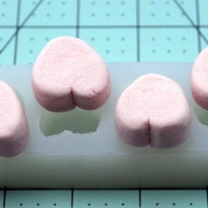 May include: Four light pink, heart-shaped clay pieces in a white silicone mold. The clay hearts have a small indentation at the bottom. The mold is on a teal cutting mat.