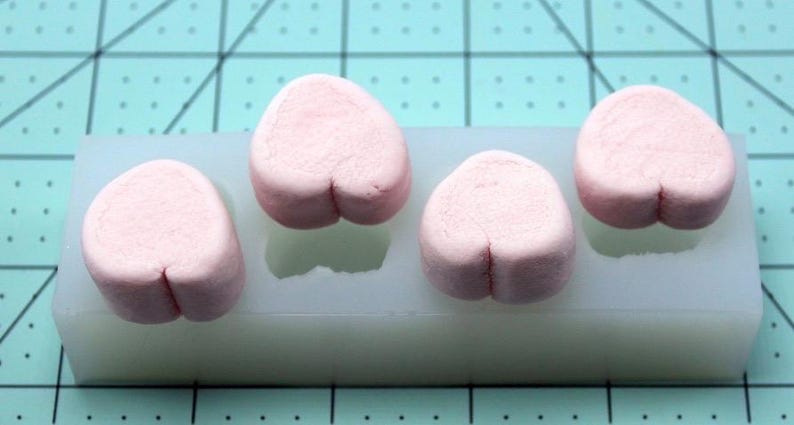 May include: Four light pink, heart-shaped clay pieces in a white silicone mold. The clay hearts have a small indentation at the bottom. The mold is on a teal cutting mat.