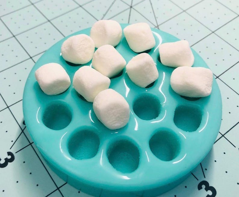 May include: A teal silicone mold with circular indentations, filled with white marshmallows. The mold is round and sits on a light blue surface with a grid pattern. The marshmallows are fluffy and evenly spaced.