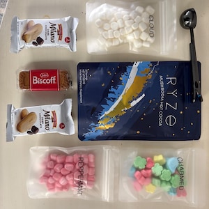 May include: An assortment of treats and ingredients for hot cocoa. Includes two Milano chocolate cookies, a Biscoff cookie, a packet of mushroom hot cocoa, marshmallows, peppermint candies, and flower-shaped candies. A black scoop is also included.