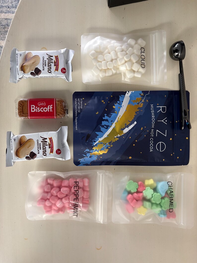 May include: An assortment of treats and ingredients for hot cocoa. Includes two Milano chocolate cookies, a Biscoff cookie, a packet of mushroom hot cocoa, marshmallows, peppermint candies, and flower-shaped candies. A black scoop is also included.