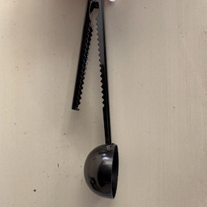 May include: A black coffee scoop with a serrated handle and a half-sphere scoop. The handle is long and slender, and the scoop is a deep, rounded shape. The scoop is made of metal and has a matte finish.