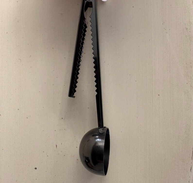 May include: A black coffee scoop with a serrated handle and a half-sphere scoop. The handle is long and slender, and the scoop is a deep, rounded shape. The scoop is made of metal and has a matte finish.